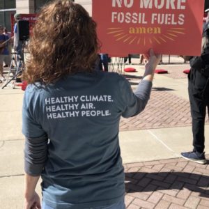 A photo from a previous NCUC carbon plan hearing shows a woman holding a sign that says "no more fossil fuels amen"